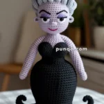 A crocheted Ursula The Sea Witch (from Little Mermaid) doll with lavender hair, bold black eyebrows, purple eyelids, a red smile, and a black dress, displayed on a table with a small shell nearby.