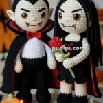 Two crocheted vampire dolls standing on a table surrounded by small decorative pumpkins and lit candles, with one doll holding a red rose.