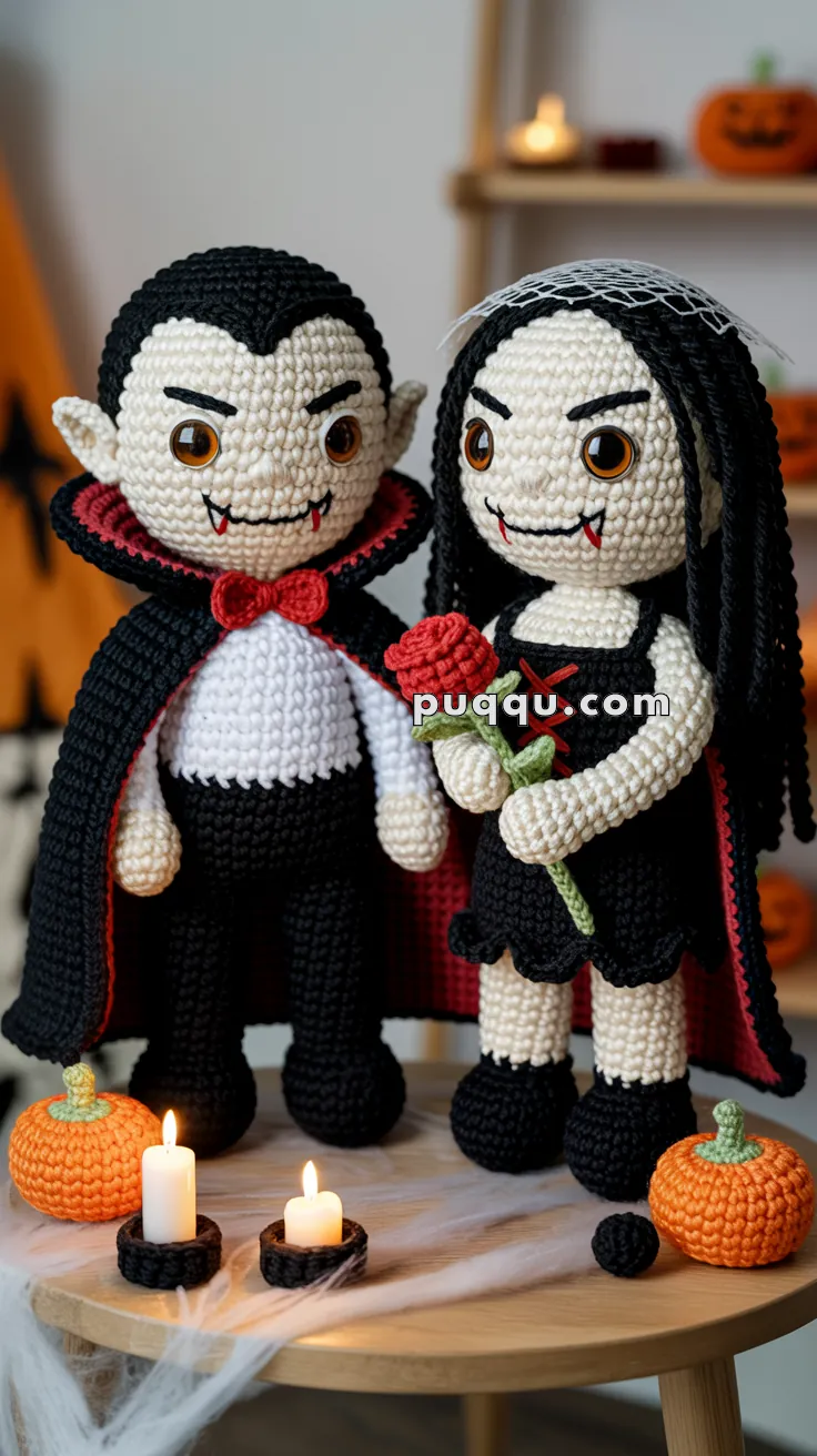 Two crocheted vampire dolls standing on a table surrounded by small decorative pumpkins and lit candles, with one doll holding a red rose.