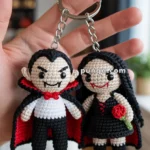 Two crocheted vampire-themed keychains, held in a hand, featuring one male and one female figure with black and red capes; the female is holding a red rose.