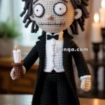 Crochet doll with black hair and wide eyes, dressed in a black tuxedo with a white shirt and bow tie, holding a crocheted candle.
