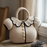 A crocheted handbag shaped like a blouse with scalloped collar and short sleeves, featuring black trim and buttons, placed on a wooden table beside a sprig of lavender.