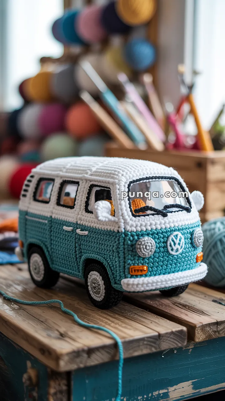 A crocheted model of a classic Volkswagen van, featuring a blue and white color scheme and intricate detailing, placed on a wooden surface with colorful yarn and crochet hooks in the background.