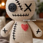 A crocheted voodoo doll with beige yarn, featuring one button eye, black stitched mouth and accents, and a red heart on its chest.