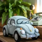 A crocheted model of a vintage car with a "MY RIDE" license plate, placed on a wooden table with a plant and rotary phone in the background.
