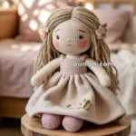 A crocheted doll with long blonde hair and a pink dress, embellished with floral embroidery, sitting on a wooden stool in a softly lit room.