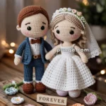 Crocheted wedding couple dolls in a suit and wedding dress with a 'FOREVER' sign and crocheted flowers on a wooden table.