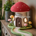 A crocheted mushroom house with a red cap sits on a wooden table, featuring a "Welcome" sign above the door and glowing windows. The house is surrounded by crocheted plants and a pathway, with a lamp and plant in the background.