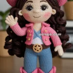 A crocheted doll wearing a pink cowboy hat, pink jacket, blue pants, and pink boots, with a golden sheriff star belt buckle, standing on a wooden surface.