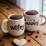 Two crocheted mugs labeled 'wifey' and 'hubby' sit on a wooden table with a crocheted heart and coffee beans scattered around.