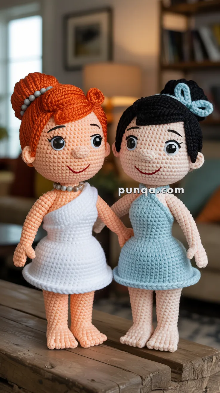 Two crocheted dolls standing on a wooden surface; one with orange hair styled in a bun and wearing a white dress, and the other with black hair in a bun and wearing a blue dress. Both are smiling and holding hands, with a home interior setting in the background.