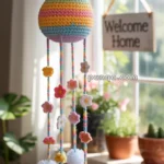 A colorful crocheted mobile featuring a rainbow-striped sphere with strings adorned with small flowers and white pom-poms, hanging near a window beside a 'Welcome Home' sign and potted plants.