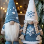 Two crocheted gnome plushies with tall hats are displayed on a wooden table. The gnome on the left has a blue hat with white stars and a white beard, while the gnome on the right has a white hat with a blue snowflake and braided hair. The background is decorated with pine branches and warm string lights, creating a cozy winter atmosphere.