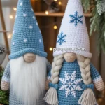 Two crocheted gnome figures, one with a blue hat and long white beard, and the other with a white hat and braided hair, both decorated with snowflake motifs, stand side by side against a festive, warmly lit backdrop.