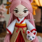 Crocheted doll with pink hair styled in two buns, wearing a traditional outfit with red and beige colors, and holding a small fox mask, with an Asian-inspired interior setting in the background.
