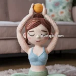 A crocheted doll in a yoga pose, sitting cross-legged on a fluffy rug, with arms raised holding a small orange ball, wearing a blue top and green pants.