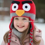 A smiling child wearing a crocheted red hat resembling a character with eyes and a beak, featuring long red and gray braided tassels, standing in a snowy outdoor setting.