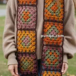A person wearing a beige sweater holds a colorful crocheted granny square scarf with squares featuring orange, green, pink, and brown yarns.