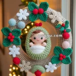 A crocheted Christmas wreath with green leaves, red berries, white flowers, and a central figure of a baby in a swaddle, hanging near a window with festive lights in the background.