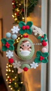 A crocheted Christmas wreath with green leaves, red berries, white flowers, and a central figure of a baby in a swaddle, hanging near a window with festive lights in the background.