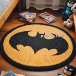 A circular crocheted rug with a black Batman logo on a yellow background is placed on a wooden floor, surrounded by children's books and a Batman plush toy.