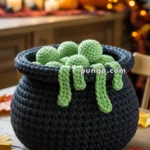 A crocheted black cauldron filled with green yarn balls and green yarn tendrils, placed on a wooden table with autumn decorations and a blurred background featuring candles and a pumpkin.