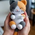A person holding a small crocheted cat doll with gray, orange, and white patches, featuring a smiling face and closed eyes, positioned in a cozy indoor setting.