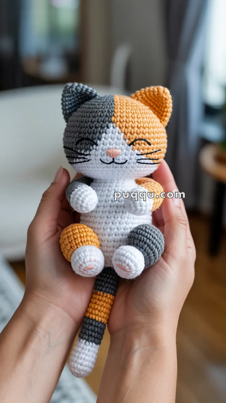 A person holding a small crocheted cat doll with gray, orange, and white patches, featuring a smiling face and closed eyes, positioned in a cozy indoor setting.