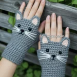 A pair of crocheted gray fingerless gloves designed to look like cat faces, with ears, whiskers, and button eyes, worn on a person's hands against a background of green leaves and wooden fencing.