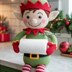A crocheted elf doll wearing a green outfit with a red collar and hat, holding a roll of paper towels, sits on a kitchen countertop decorated with festive holiday ornaments.