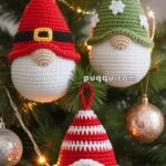 Three crocheted gnome ornaments hanging on a Christmas tree, each with a distinctive hat: one red with a belt buckle, one green with white stars, and one red and white striped.