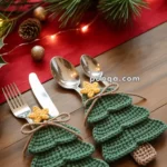 Table setting with forks and spoons placed on knitted Christmas tree-shaped holders, alongside festive decorations including pinecones and holly on a red runner.
