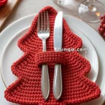 A festive table setting featuring a red crocheted Christmas tree-shaped napkin holding a fork and knife, placed on a white plate, with a glass of red wine and a water glass in the background.
