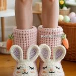 Pink knitted bunny slippers with floppy ears and carrot accents on a wooden floor.