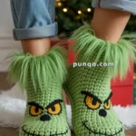 Green Grinch-themed slippers with fuzzy tops, featuring a mischievous face design, worn by a person in rolled-up jeans, standing near a Christmas tree.