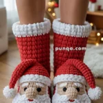 Person wearing knitted Santa Claus themed slippers with red and white colors, standing on a wooden floor with blurred Christmas decorations in the background.