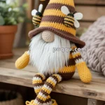 A crocheted gnome doll with a long beard, wearing a yellow and brown striped hat adorned with crochet bees, sitting on a wooden surface.