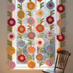 A window decorated with a crochet curtain featuring colorful flowers and green leaves, with a wooden rocking chair placed nearby on a wooden floor.