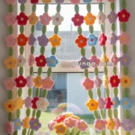 Crocheted flower curtain with colorful flowers and green stems draped over a window, with a crocheted flower pot on the windowsill.
