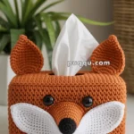 A crocheted tissue box cover designed to look like a fox, featuring orange yarn for the body, white yarn for the cheeks, black buttons for eyes, and pointed ears. A tissue peeks out from the top. A plant and a wicker basket are blurred in the background.