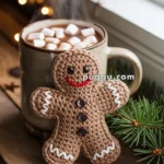 A crocheted gingerbread man decoration rests next to a steaming mug of hot chocolate topped with marshmallows, on a wooden surface adorned with festive stars and evergreen branches.