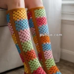 Person wearing knee-high crochet socks with colorful granny square patterns, sitting on an armchair in a room with light-colored walls and floor.