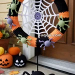 A Halloween-themed crochet wreath featuring a spider web and several colorful crocheted spiders in black, green, and purple. Below the wreath, a large crocheted black spider hangs, with smiling features. The wreath is displayed on a front porch with crocheted pumpkins in the background and autumn leaves scattered on the ground.