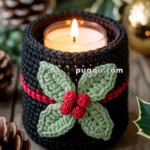 A candle in a glass holder wrapped in a knitted black cover with red accents, decorated with crocheted green leaves and red berries, surrounded by pine cones and festive holiday decorations on a wooden surface.