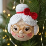 A crocheted ornament of an elderly woman with glasses and a red bow, hanging on a Christmas tree branch with lights.