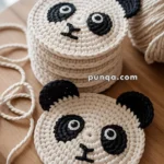 Crochet coasters designed to resemble panda faces, featuring black ears and eye patches, displayed on a wooden surface with yarn visible in the background.