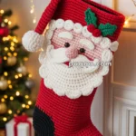 A handmade crochet Christmas stocking featuring a Santa Claus design, hanging on a fireplace mantel with decorative greenery and fairy lights. In the background, there is a Christmas tree adorned with ornaments and a few wrapped presents nearby.