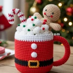 A crocheted mug designed to look like a Santa suit, filled with crocheted whipped cream, colorful sprinkles, a candy cane, and a smiling gingerbread man, set on a wooden table with a Christmas tree in the background.