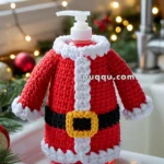 A soap dispenser dressed in a crocheted cover resembling a Santa Claus outfit, featuring red fabric with white trim and a yellow belt buckle, set on a kitchen counter with festive decorations and lights in the background.