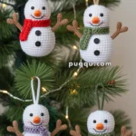 Crocheted snowman ornaments with carrot noses, button eyes, and twig arms, wearing scarves in red, green, purple, and also green, are hanging on a Christmas tree adorned with lights.